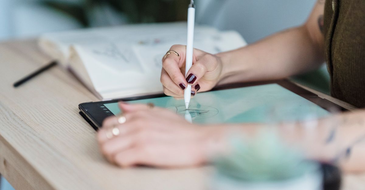 Crop unrecognizable female designer using tablet and stylus for drawing sketches on wooden table